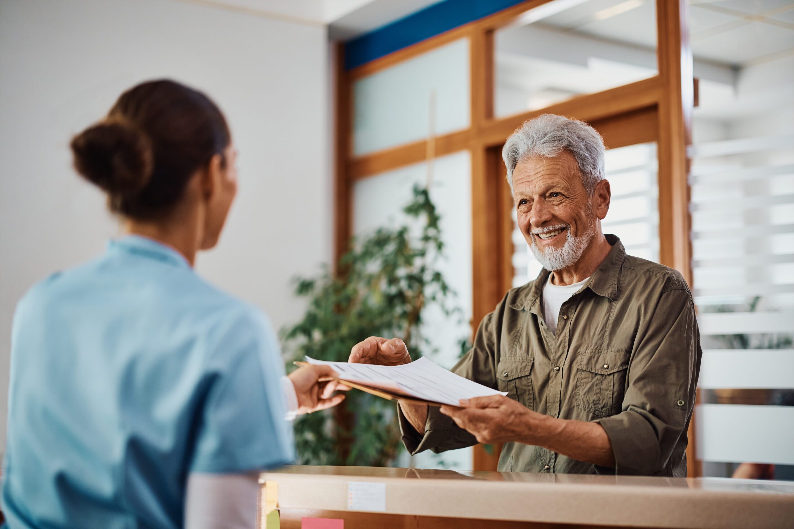 Happy senior man receiving his medical documents from nurse at reception desk at doctor's office.